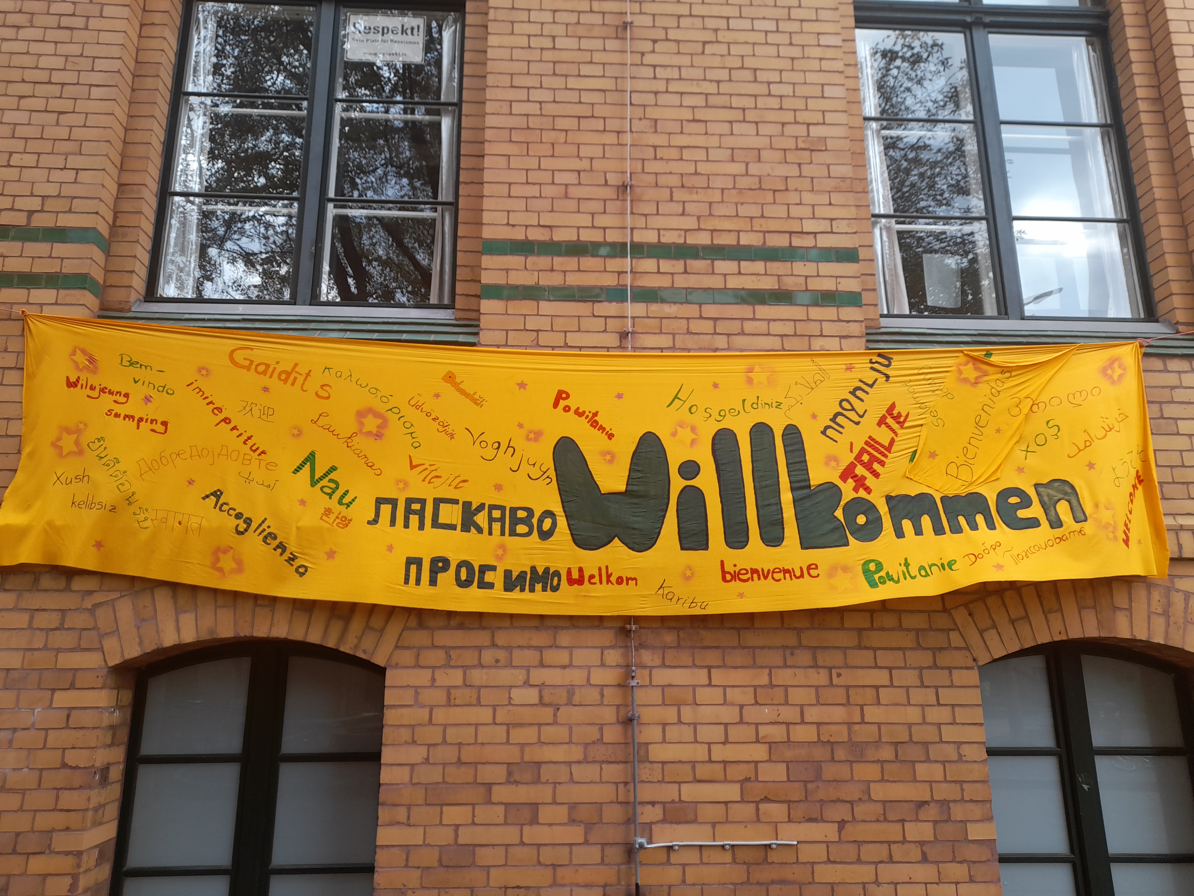 Zu sehen ist ein gelbes Banner an dem Gebäude der Reinhold-Burger-Schule. Auf dem Banner steht "Willkommen" auf verschiedenen Sprachen.