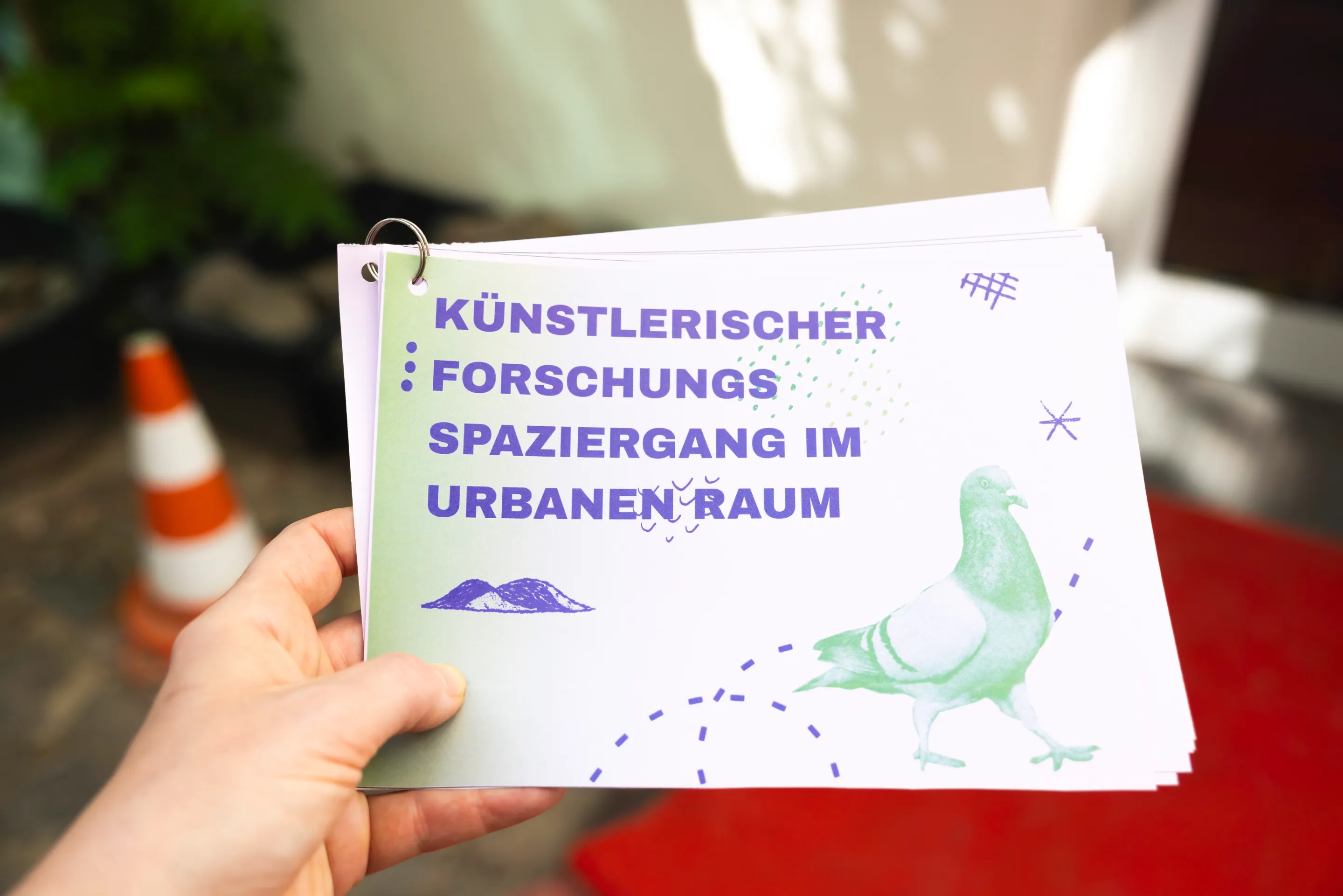 Zu sehen ist, eine Hand, die ein Booklet mit dem Schriftzug "künstlerischer Forschungsspaziergang im urbanen Raum" hält.