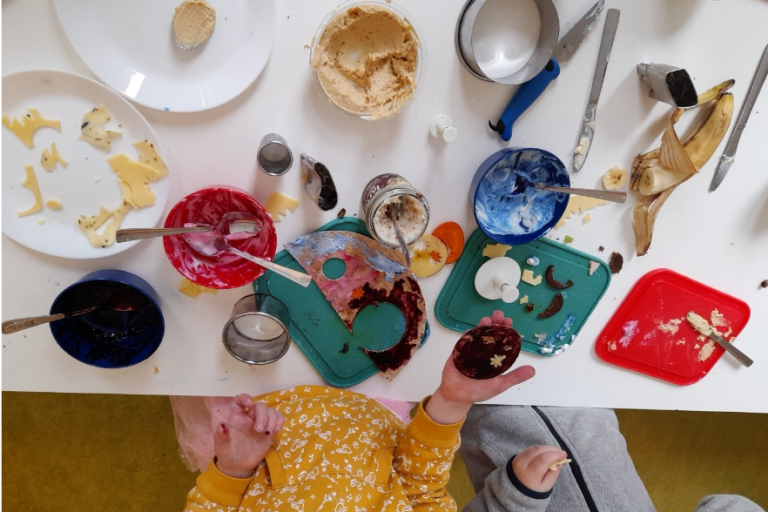 Es ist ein Tisch von oben zu sehen, an dem Kinder mit Essen experimentieren und besondere Butterbrote erstellen.